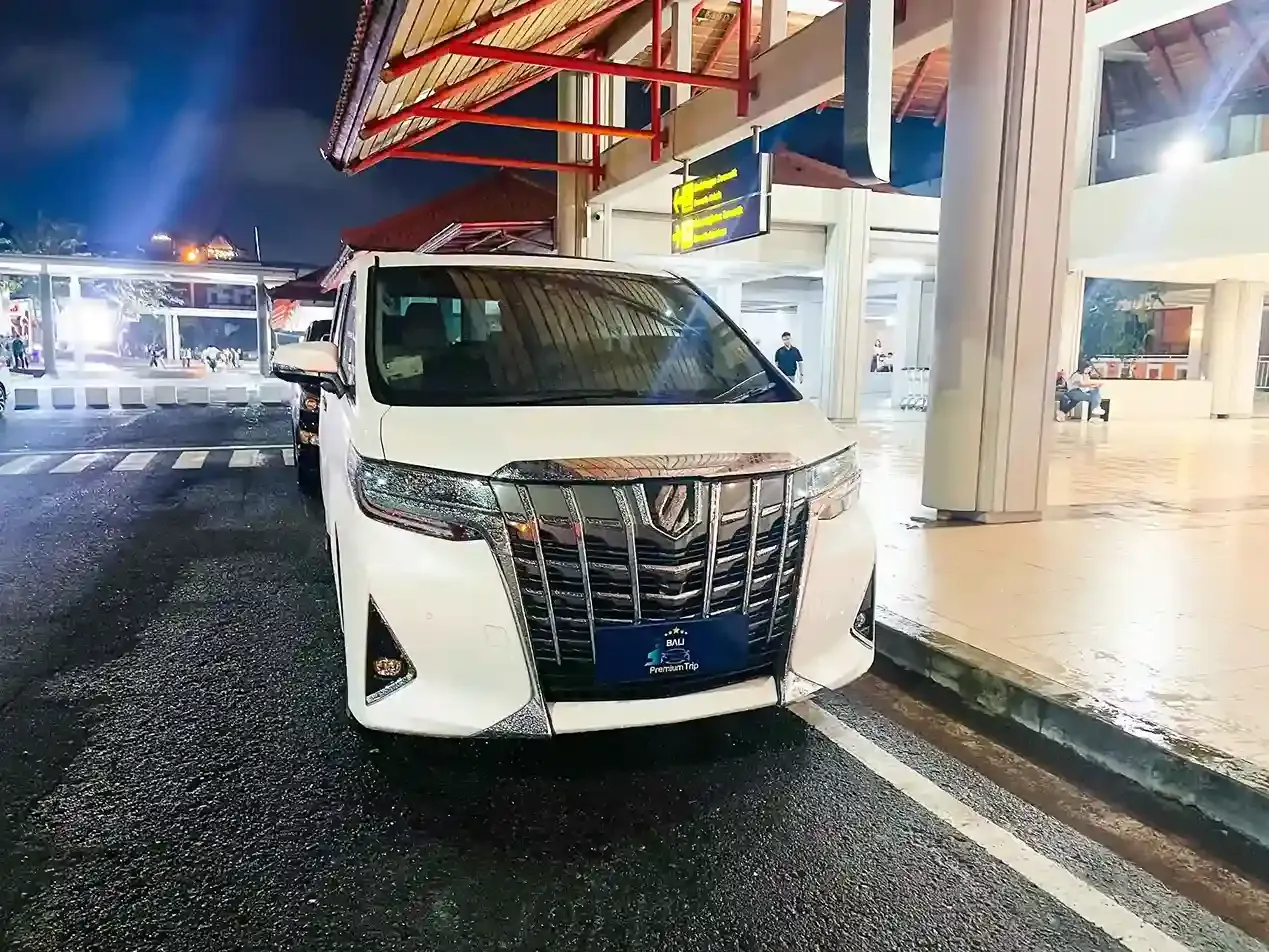 A white Toyota Alphard waiting at the Ngurah Rai terminal kerb at night