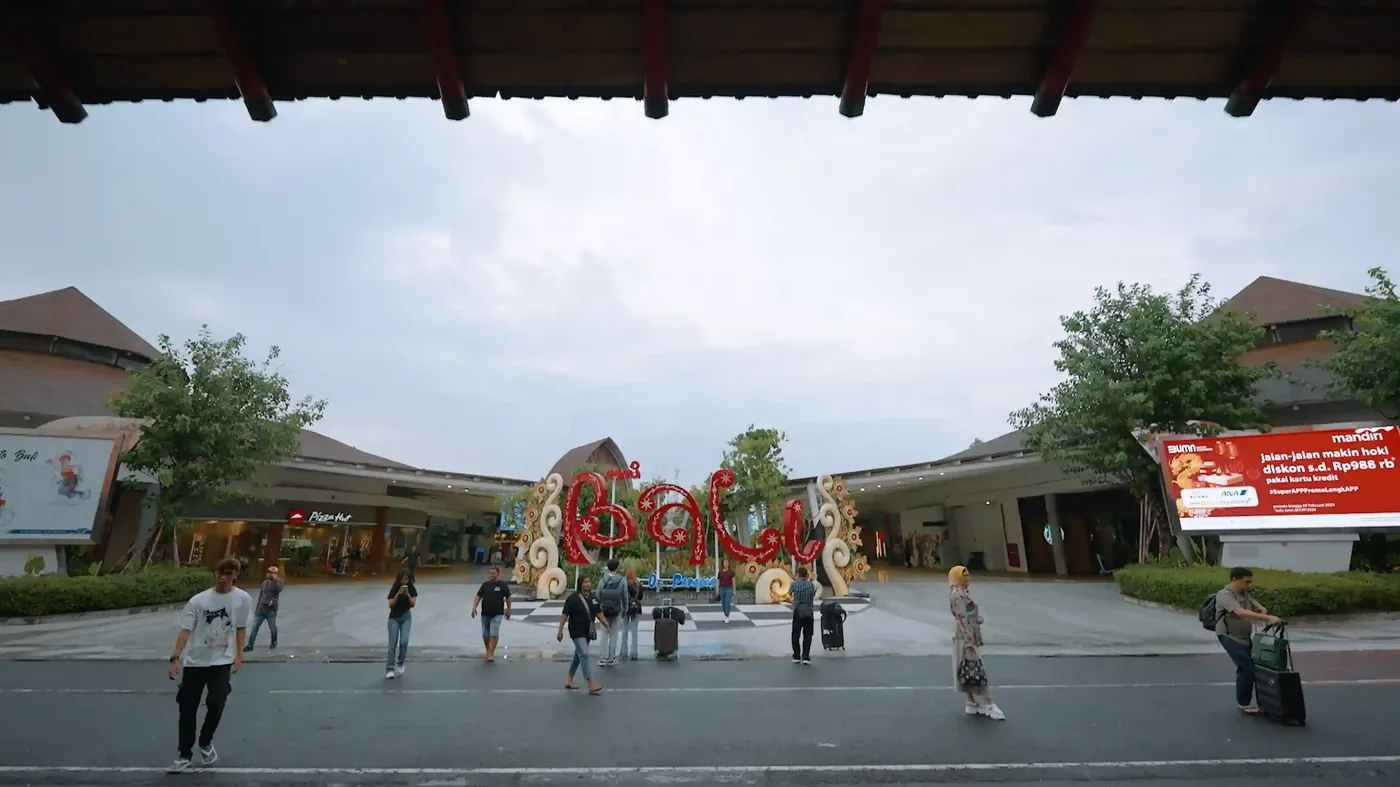 Travellers with luggage crossing the forecourt by the BALI sign at Ngurah Rai airport