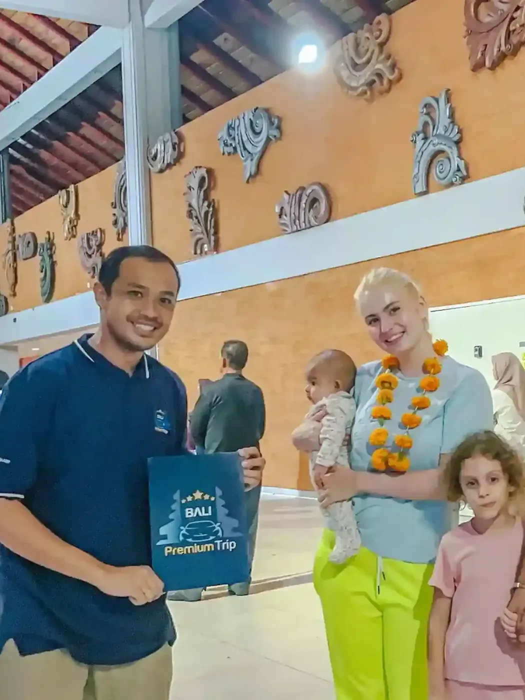 A greeter holding a Bali Premium Trip folder welcomes a family wearing flower garlands in the Ngurah Rai arrivals area