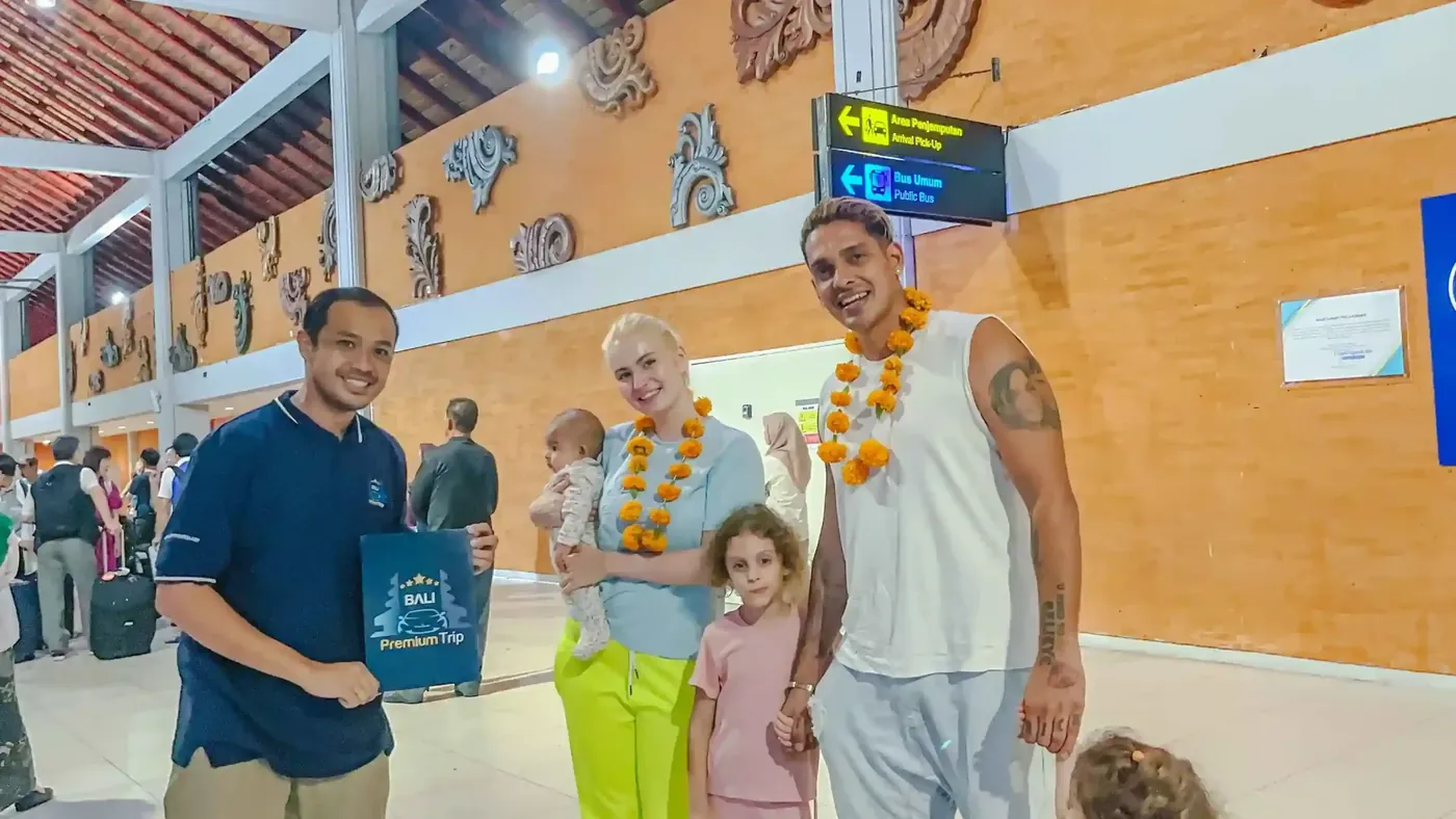 A greeter holding a Bali Premium Trip folder welcomes a family wearing flower garlands in the Ngurah Rai arrivals area